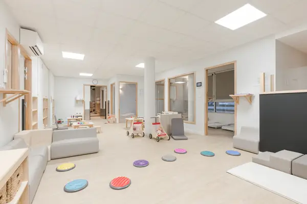 Salle de vie de la crèche Les Bébés Explorateurs Angers (49100) – disques sensoriels colorés au sol, banquettes modulaires grises, porteurs, accès aux dortoirs par ouvertures à encadrement bois et claustra bois